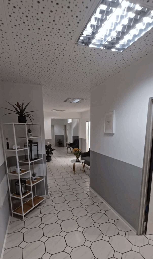 Heller Praxisflur mit Sitzbereich, Pflanzen und Regal; moderne Fliesen, klare Linien, freundliche Atmosphäre.