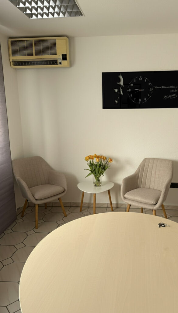 Helles Beratungszimmer mit zwei Sesseln, kleinem Tisch mit Tulpen und Wanduhr – freundliche Praxisatmosphäre.